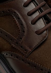 Scarpa di pelle marrone con una finitura liscia, dettagli in suede, perforazioni decorative e lacci texturizzati. Il design include cuciture a zigzag.