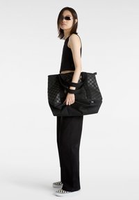Zwarte schoudertas met een geruit patroon, grote maat, stoffen textuur, dubbele handvatten en een matte afwerking. Draagt met zwarte kleding en geruite schoenen.