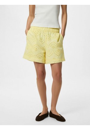 Femeie poartă pantaloni scurți galben pal, cu ochiuri, talie elastică și buzunare, asortați cu un top alb fără mâneci și balerini maro închis.