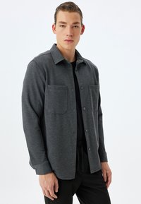 Camisa gris de botones hecha de tela texturizada, con dos bolsillos en el pecho y un corte relajado, combinada con pantalones oscuros.