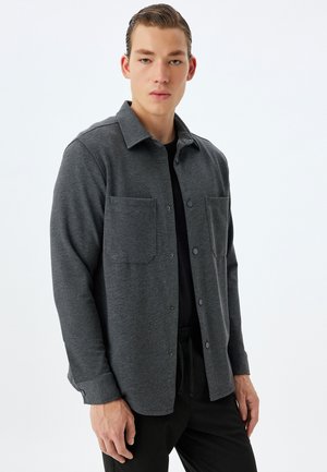 Camisa gris de botones hecha de tela texturizada, con dos bolsillos en el pecho y un corte relajado, combinada con pantalones oscuros.