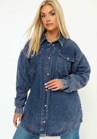 Camicia denim oversize di colore blu scuro con due tasche sul petto, chiusura a bottoni e orlo arrotondato. Presenta maniche lunghe e una vestibilità rilassata.