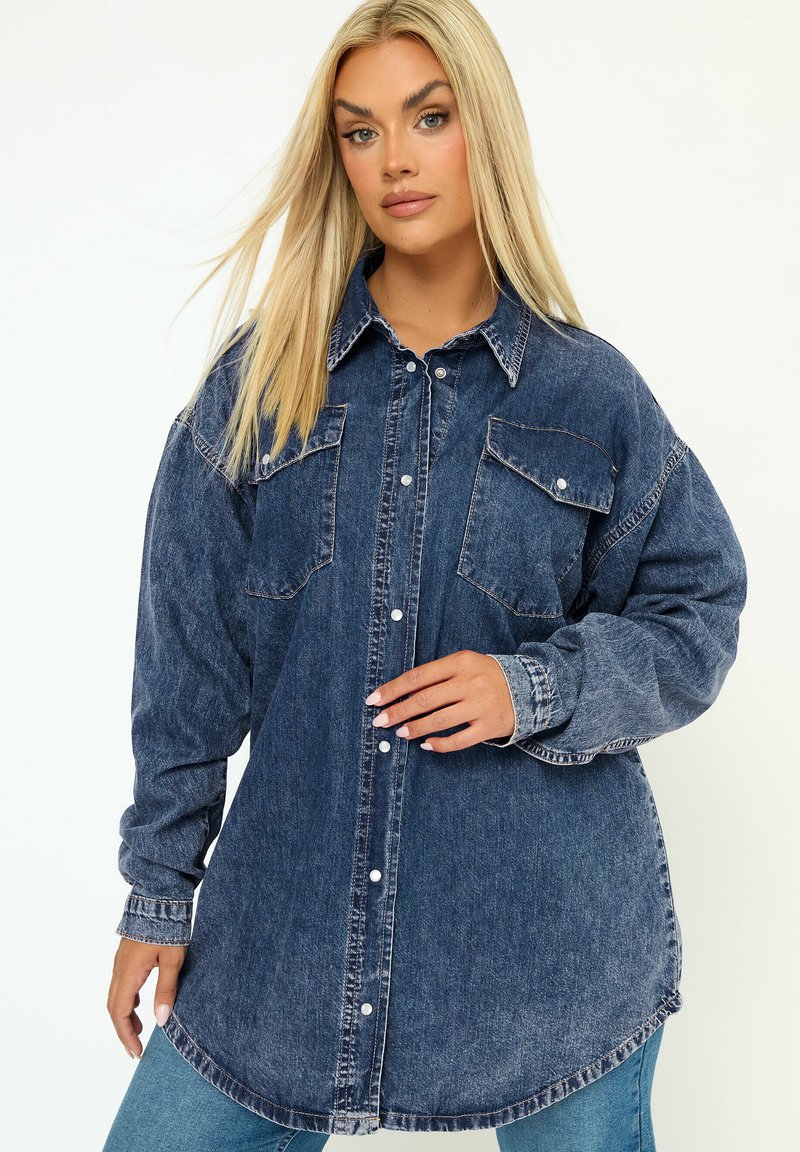 Camicia denim oversize di colore blu scuro con due tasche sul petto, chiusura a bottoni e orlo arrotondato. Presenta maniche lunghe e una vestibilità rilassata.