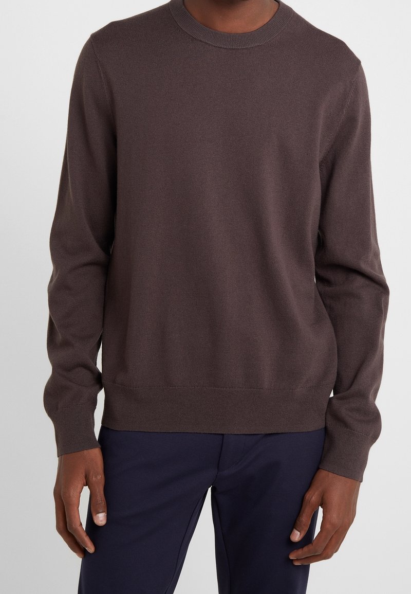 Homme portant un pull uni marron foncé à manches longues et col rond, ainsi qu’un pantalon bleu foncé, sur un fond clair.