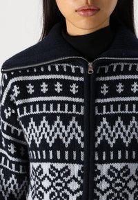 Pletený kardigan v námořnické modři a bílé, s geometrickým vzorem, vysokým límcem a stříbrným zipem. Textura je silná a vlněná.