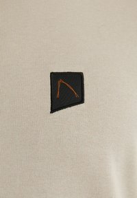 Beige fabric features a black rectangular patch with an orange geometric design stitched on it. The texture is smooth and fitted.