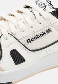 Baskets Reebok avec une tige en cuir principalement blanche, des accents noirs, des détails en caoutchouc texturé et des lacets beiges.