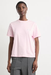 Pink cotton t-shirt with short sleeves, a round neckline, and a small logo on the left chest. Paired with grey, wide-leg trousers.