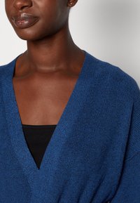Blå stickad cardigan med djup V-ringning. Tillverkad av texturerat tyg, har den avslappnade ärmar och passar bra med en svart topp under.