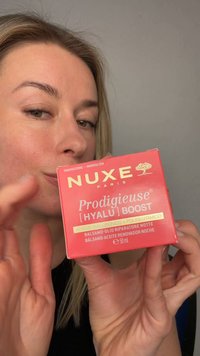 Vrouw houdt een rode doos van NUXE Prodigieuse Hyalu Boost nacht herstel balsem dicht bij haar gezicht tegen een effen achtergrond.