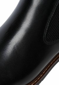 Bota de cuero negro con una textura suave, paneles laterales elásticos y un ribete marrón en contraste a lo largo de la suela. Vista en primer plano centrada en los detalles.