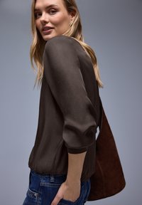 Blouse marron texturée avec poignets ajustés, ourlet légèrement élastiqué, associée à un jean en denim bleu et un sac à main en daim marron.