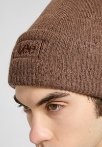 Cappello beanie marrone lavorato a maglia con texture a coste e un'etichetta rettangolare. Il tessuto appare morbido e caldo, adatto per il tempo freddo.