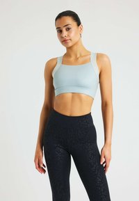 Vrouw staat, gekleed in een lichtblauwe sport-bh met dikke bandjes en hooggeplaatste zwarte leggings met een subtiel luipaardmotief.