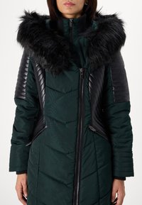 Green puffer coat with a black faux fur hood. Features quilted shoulder accents and zippered side pockets. Clean lines and modern design.
