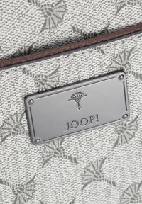 Szary, teksturowany materiał z kwiatowym wzorem oraz srebrna metalowa blaszka z wyrytą stylizowaną kwiatu i napisem "JOOP!".