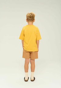 Gul bomulds T-shirt med korte ærmer, parret med brune shorts. Drengen har lyse sokker og brune sandaler på. Enkel design.