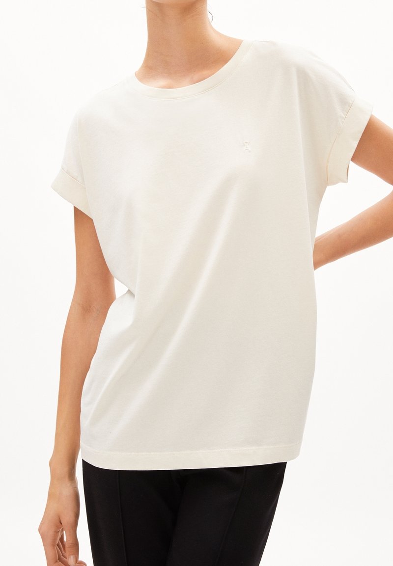 Femme portant un t-shirt ample à manches courtes couleur écru avec des manches retroussées et un pantalon noir, posant avec une main sur la hanche.