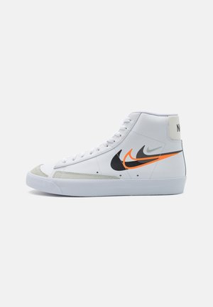 Białe wysokie buty sportowe Nike z wielowarstwowymi logo Swoosh w kolorach czarnym, pomarańczowym i szarym po boku oraz szarymi zamszowymi wstawkami na nosku.