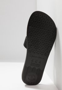 Semelle noire en caoutchouc d'une tong avec bande de roulement texturée et marquages de taille et de marque, debout contre un mur d’angle blanc.