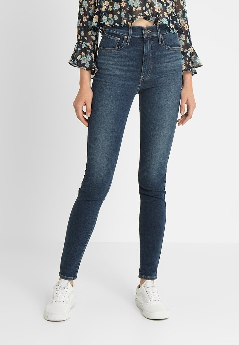 Femme portant un jean skinny bleu foncé, un chemisier court à motifs floraux et des baskets blanches, debout devant un fond clair uni.
