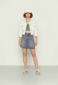 Jaqueta de ganga branca, top cropped cinza com padrão e saia de ganga azul com bainha desfiada. Um boné branco e sandálias completam o visual casual.