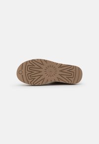 Semelle de chaussure marron clair avec un motif de bande de roulement en forme d'éclat de soleil géométrique et un petit détail circulaire près de la zone des orteils sur un fond uni.