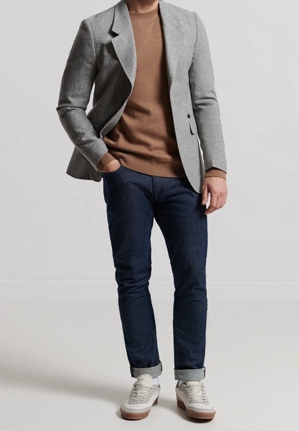 Homme portant un pull marron, un blazer gris, un jean bleu foncé retroussé et des baskets blanches, debout avec une main dans la poche.