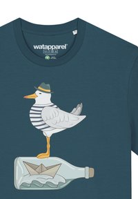 Cyan t-shirt met een afbeelding van een meeuw in een gestreept shirt en groene hoed, die op een fles staat met een papieren bootje erin.