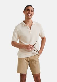 Polo-shirt à manches courtes de couleur crème avec un tissage texturé, doté un col à deux boutons, associé à un short beige clair.