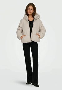 Kvinde iført en beige dunjakke over en sort knap-top, sorte flagrende bukser og sorte spidse sko, stående med hænderne i lommerne.