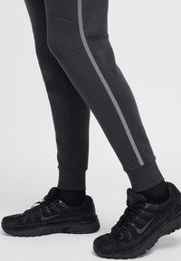 Melnas sporta bikses ar pelēku sānu svītru un pieguļošiem manžetēm, kopā ar melniem Nike sporta apaviem ar teksturētu virsmu un mīkstinātām zolēm.