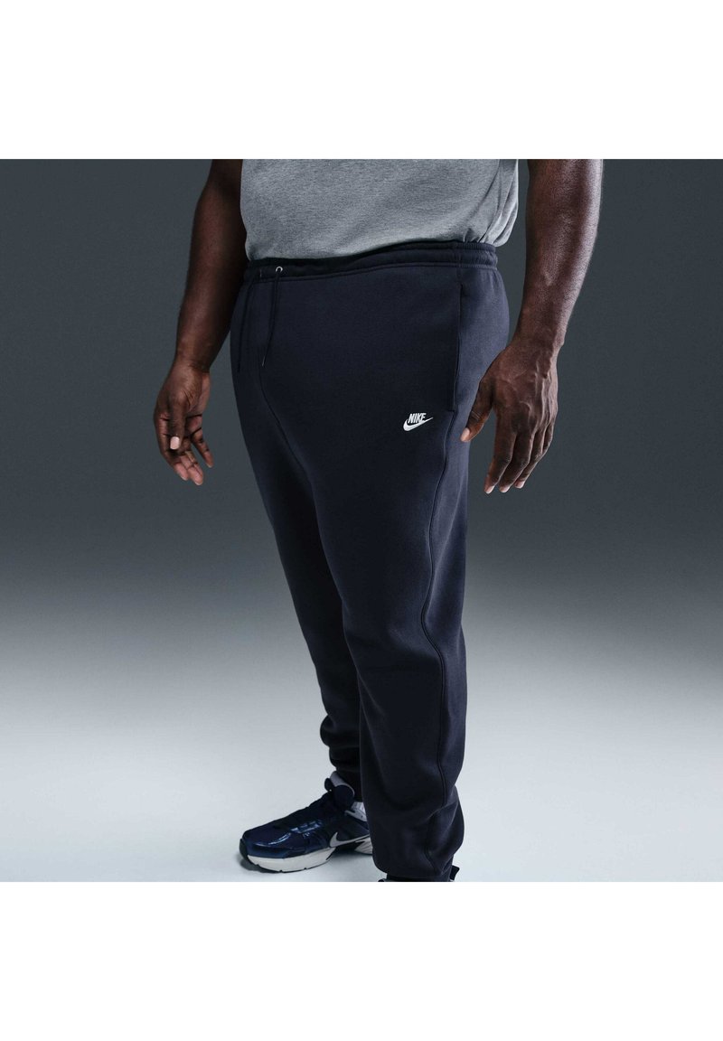 Persona con pantaloni da tuta Nike neri, maglietta grigia e scarpe da ginnastica nere che sta in piedi davanti a uno sfondo grigio semplice. È visibile solo la parte inferiore del corpo.
