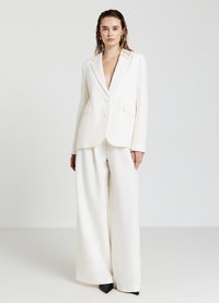 Blazer bianco su misura con rever a punta, chiusura con un solo bottone e due tasche frontali; abbinato a pantaloni bianchi a gamba larga e tacchi neri.