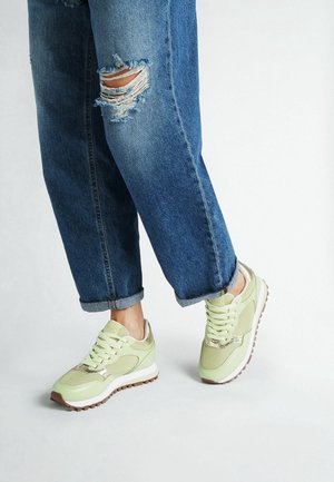 Persona che indossa jeans blu strappati e risvoltati e sneaker in mesh verde chiaro con suole bianche e battistrada marroni, in piedi su pavimento grigio chiaro.