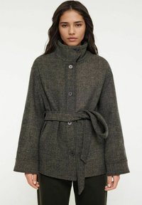 Mörkgrön rutig ullkappa med hög krage, framknappar och ett knytbälte. Har vida ärmar och texturerad tyg.