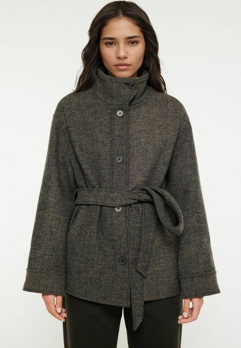 Mörkgrön rutig ullkappa med hög krage, framknappar och ett knytbälte. Har vida ärmar och texturerad tyg.