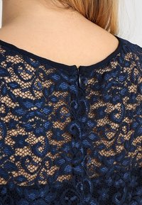 Haut en dentelle bleu marine avec un motif floral, dos texturé avec une fermeture en forme de clé et un col noir lisse.