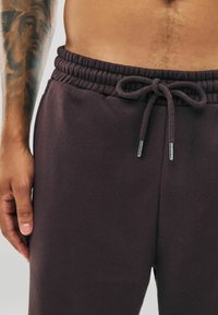 Dunkelviolette Sweatpants mit elastischem Bund, der über eine Kordelzugverschluss verfügt und silberne Akzente aufweist. Glatte Textur.