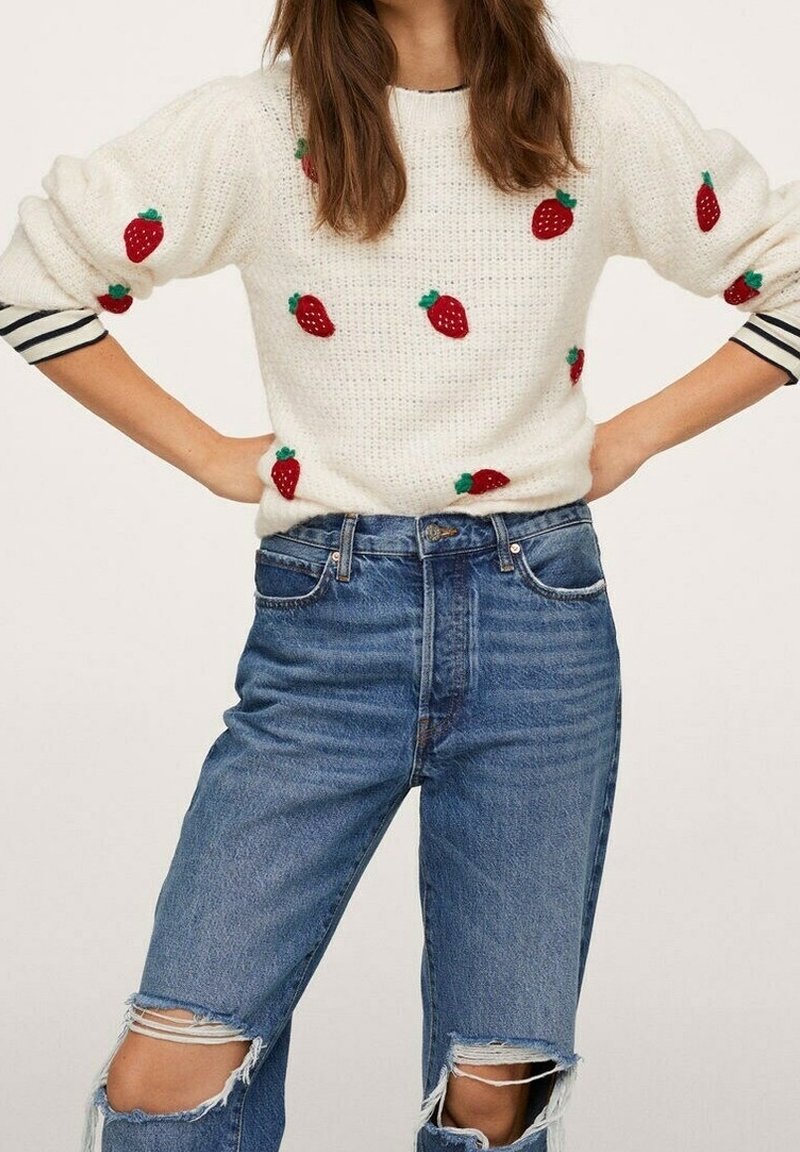 Femme portant un pull en maille blanc avec des motifs de fraises rouges sur une chemise à manches longues rayée, associé à un short en jean bleu déchiré.