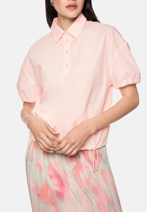 Femme portant un chemisier rose clair à manches courtes bouffantes et une jupe plissée pastel avec des motifs roses et verts.