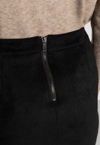 Jupe en suede noire au texture lisse, avec une fermeture à glissière sur le côté et une coupe ajustée, associée à un haut en tricot beige.