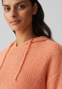 Felpa con cappuccio di colore pesco in maglia, con tessuto texturizzato, dotata di cappuccio regolabile e motivo a costine nel corpo. Vista ravvicinata del collo.
