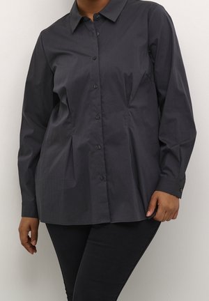 Personne portant une chemise à manches longues, boutonnée, gris foncé avec un col, associée à un pantalon noir sur un fond uni.