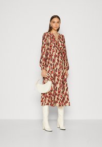 Abito lungo con motivo in beige, con forme astratte rosse e marroni, maniche lunghe e scollatura a V profonda. Abbinato a stivali bianchi fino al ginocchio e una borsa bianca.