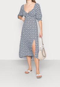 Květinové midi šaty v námořnické modři s bílými a modrými vzory; mají lodičkový výstřih, nabírané rukávy a boční rozparek. Spárováno s lehkou taškou.