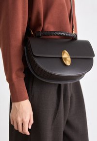Sac bandoulière en cuir noir avec poignée tressée et fermoir ovale doré. Forme arrondie, texture lisse et détails de design minimalistes.