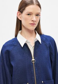 Τζιν μπουφάν σε σκούρο μπλε χρώμα με λευκό πουκάμισο με γιακά από μέσα, διαθέτει χρυσό φερμουάρ, δύο τσέπες μπροστά και άνετη εφαρμογή.