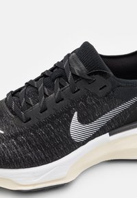 Chaussure de sport noire avec une tige en tricot texturé, des accents réfléchissants, un swoosh blanc et une semelle rembourrée avec une base de couleur crème.