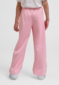 Osoba ubrana w luźne jasnoróżowe spodnie i białe trampki, stojąca tyłem do widza na jednolitym tle.